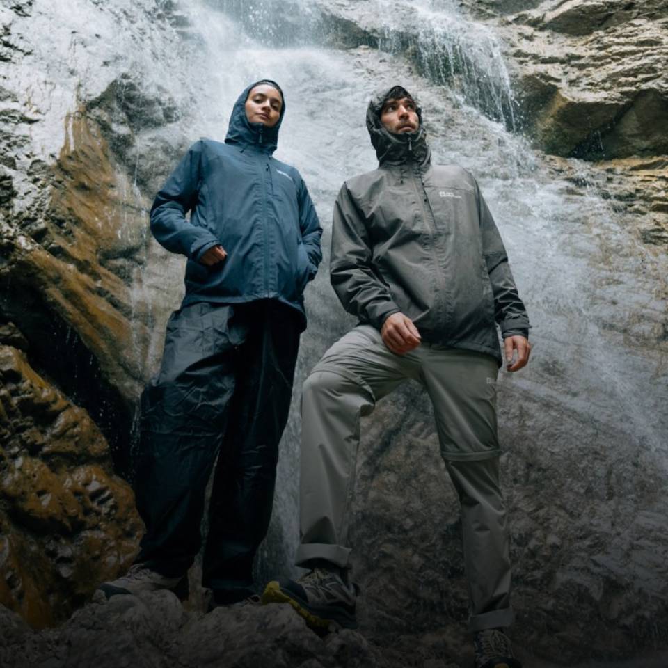 Two hikers in sporty rain jackets stand in front of a waterfall